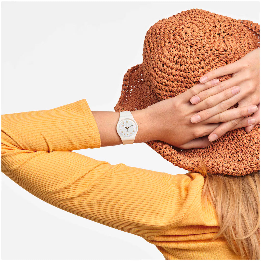 Orologio Swatch bianco indossato al polso, abbinato a una maglia gialla e a un cappello intrecciato.