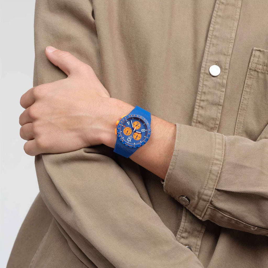 Swatch PRIMARILY BLUE SUSN419 indossato al polso con camicia beige, quadrante blu con tre contatori arancioni e cinturino blu coordinato.