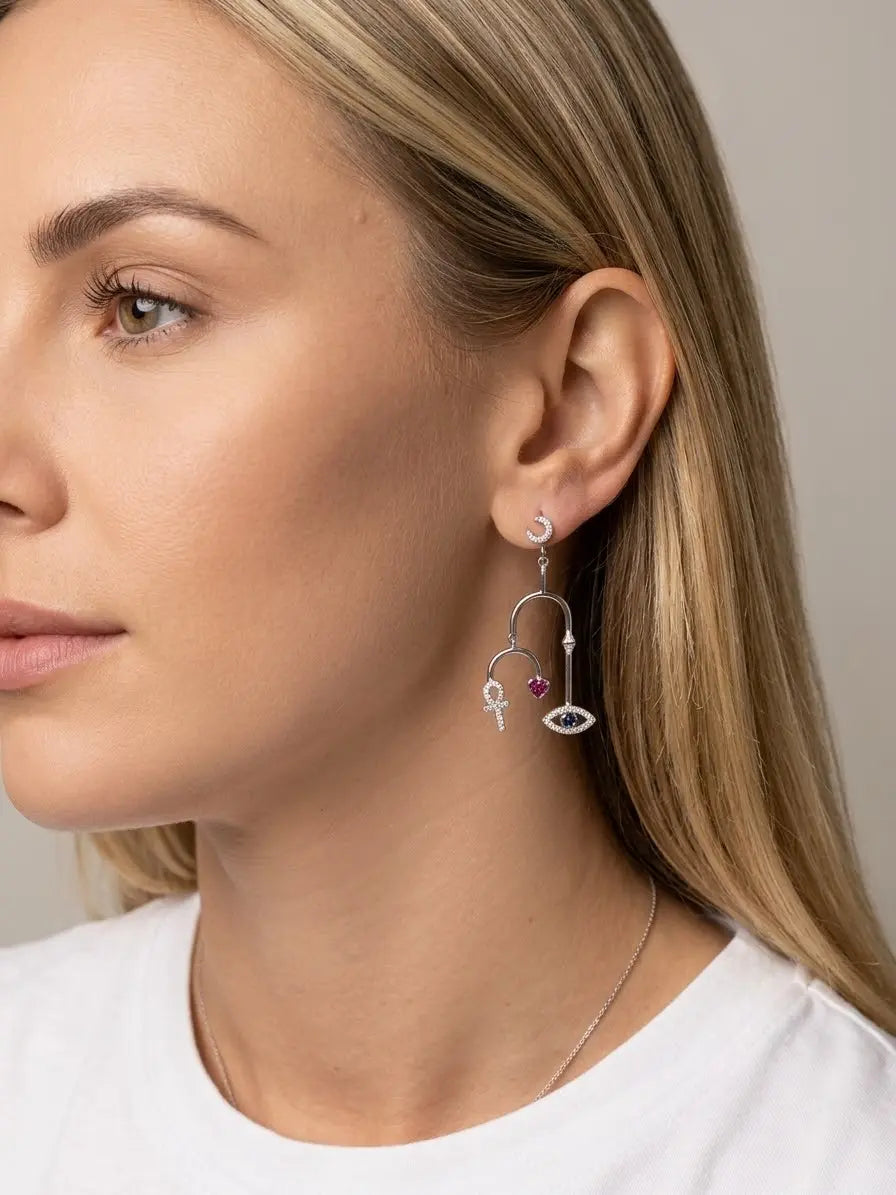 Orecchino asimmetrico OR0139 in argento 925 indossato, con struttura ad archi e pendenti a croce ansata, cuore rosa e occhio blu, CICCO Gioielleria.